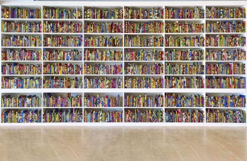 An image of books on a bookshelf that fills the entire photograph. The books are bound in African wax print fabric, printed in gold on the spines of 2,700 of the books are the names of first or second-generation immigrants to Britain.