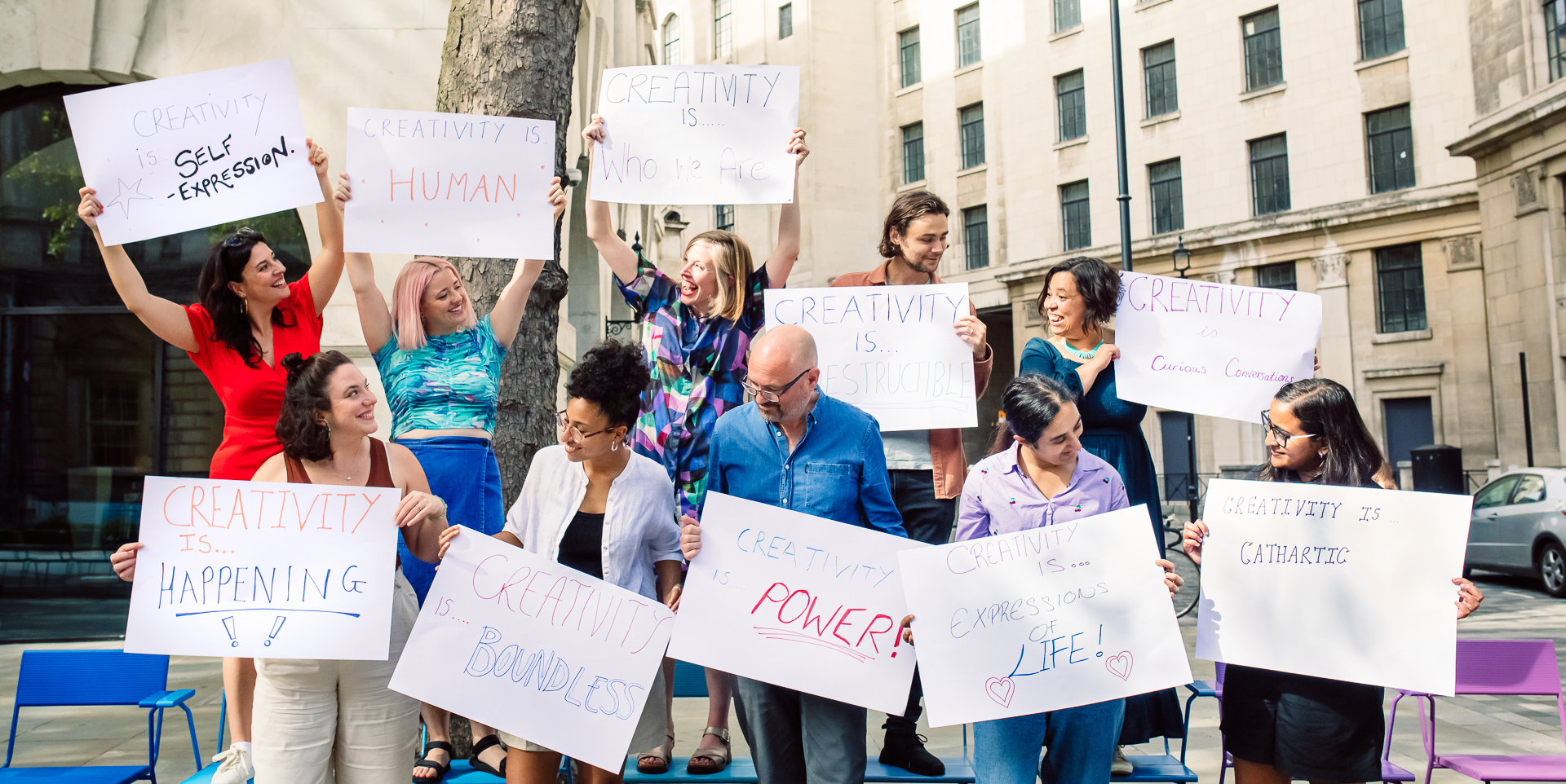 A picture of the 64 Million Artists team, standingon chairs and holidng up big 'Creativty Is...' posters