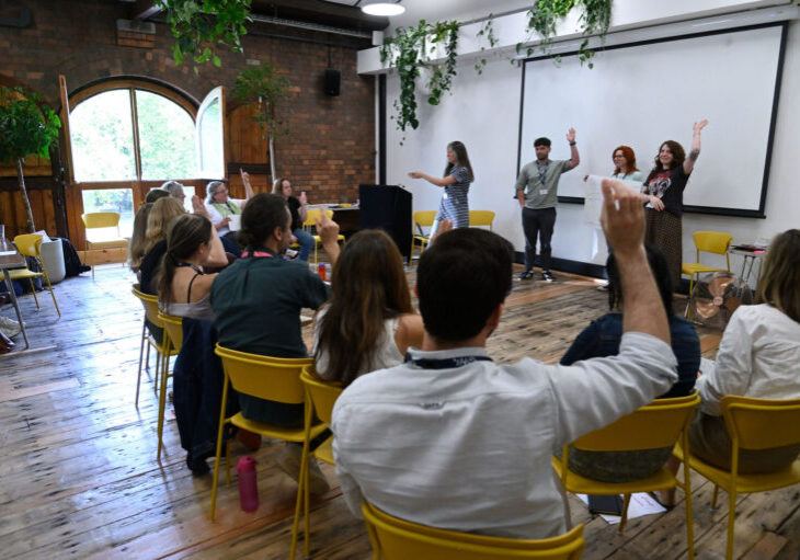 A workshop participant raising their hand during a group leadership session in a bright event room.